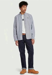 Camisa azul claro de botões com mangas longas, jeans cinzentos e botins castanhos. Apresenta uma t-shirt branca com texto preto por baixo da camisa.