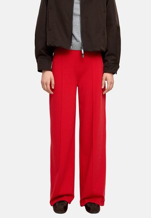 Osoba ubrana w jaskrawo czerwone spodnie o szerokich nogawkach, czarne buty, szary top i ciemnobrązową kurtkę zapinaną na zamek, stojąca na jednolitym tle.