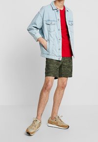 Denimjakke, lyseblå; rød t-shirt; grønne camo-shorts; lysebrune sneakers med mørkere accenter; afslappet outfit.
