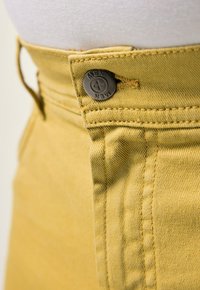 Žluté bavlněné kalhoty s kovovým knoflíkem s vyrytým nápisem "MEN". Podrobný šev podél pasu a bočních švů.