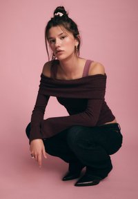 Lila, geripptes, schulterfreies Oberteil mit langen Ärmeln, kombiniert mit dunklen Jeans und spitzen schwarzen Stiefeln. Haare zu einem Dutt mit einem rosa Haargummi gebunden.