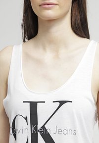 Bílý tílko s velkým černým logem "CK" a textem "Calvin Klein Jeans" pod ním, vyrobené z lehké a hladké tkaniny.