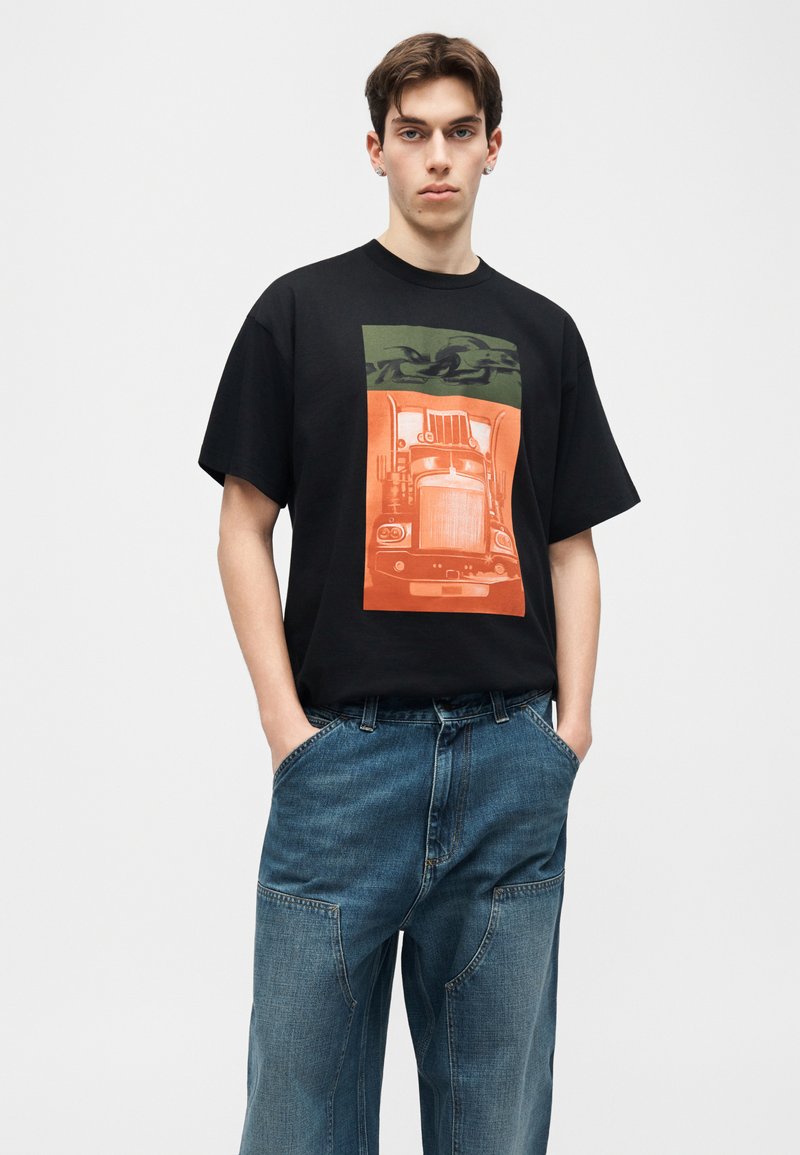 Joven con camiseta negra con un gráfico naranja y verde de un camión, combinado con vaqueros azules holgados, de pie con las manos en los bolsillos.