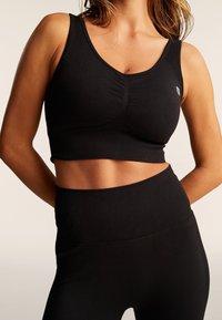 Donna che indossa un reggiseno sportivo nero aderente e leggings a vita alta, mostrando l'addome su uno sfondo neutro.