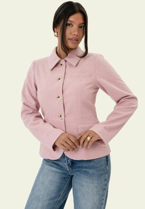Femme portant une veste rose à boutons avec col et un jean bleu, debout les mains jointes à la taille devant un fond uni.