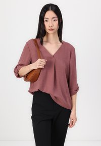 Løs-fittende bluse med dyp v-hals i teksturert mauve, kombinert med svarte bukser. Har rullet ermer og bæres med en brun crossover-veske.