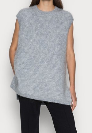 Femme portant un gilet sans manches en maille grise sur un pantalon noir, debout devant un fond blanc uni.