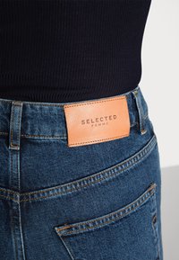 Modré denimové džíny s oranžovým šitím. Mají koženou nášivku hnědé barvy s embosovaným nápisem "SELECTED FEMME". Rovný střih a zadní kapsy jsou viditelné.