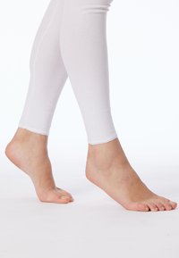 Leggings blancs ajustés en tissu lisse et extensible, se terminant juste au-dessus de la cheville, portés pieds nus.