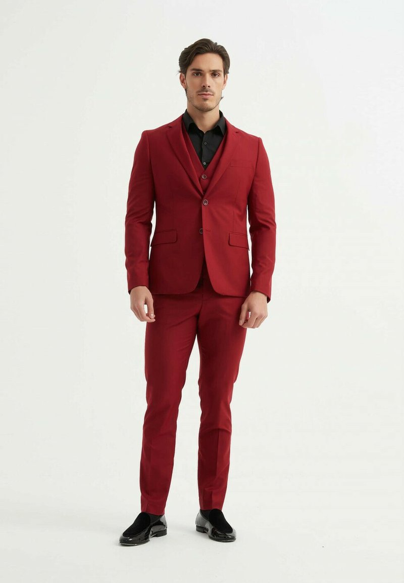 Costume rouge avec pantalon assorti, coupe slim, veste à un bouton, deux boutons, revers crantés, porté sur une chemise noire, chaussures noires brillantes.