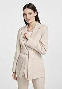 Beżowa marynarka z długimi rękawami, klapami w szpic i związanym w pasie. Charakteryzuje się minimalistycznym designem i gładkim materiałem.