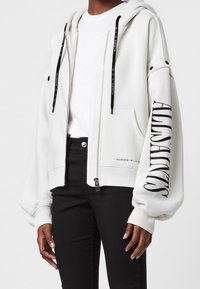 Vit zip-hoodie med en rymlig huva, svarta dragsnören och en framträdande logotyp tryckt på vänster ärm. Mjuk textur, avslappnad passform.