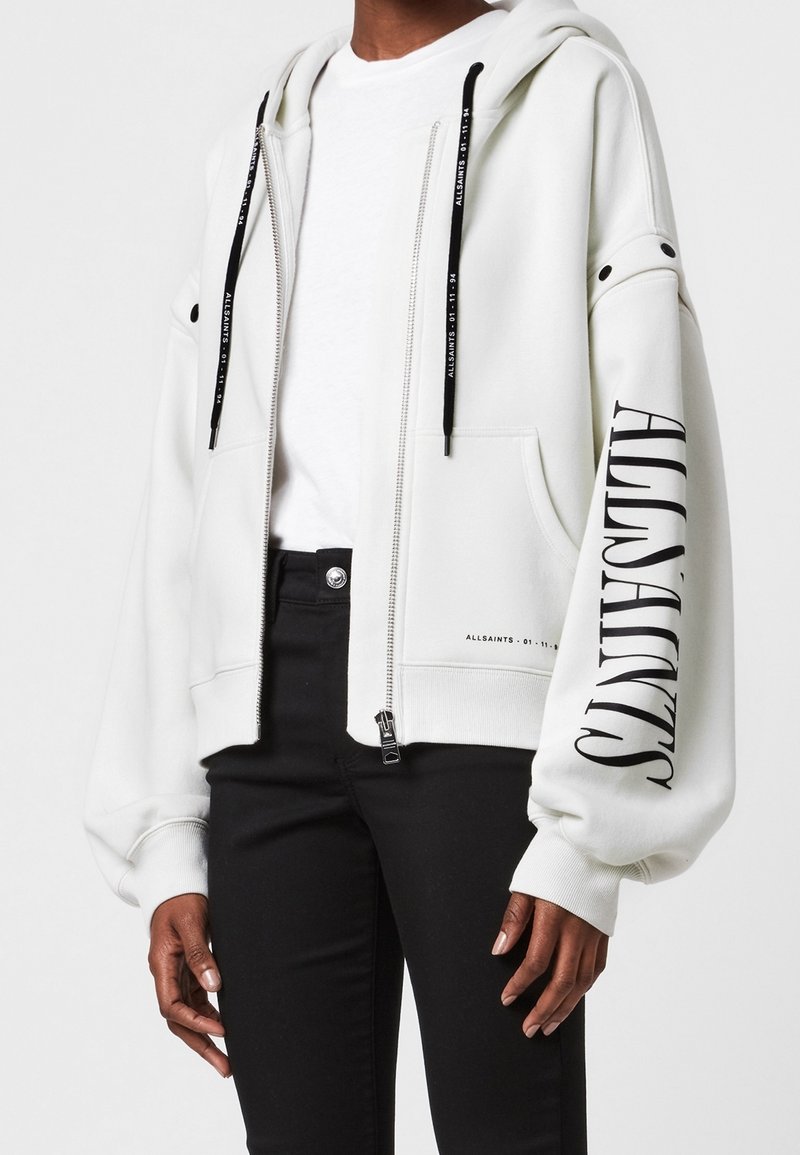 Vit zip-hoodie med en rymlig huva, svarta dragsnören och en framträdande logotyp tryckt på vänster ärm. Mjuk textur, avslappnad passform.