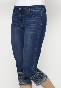 Capris en denim bleu foncé avec une coupe ajustée. Présentent des accents décoratifs avec des motifs et des franges au niveau de l'ourlet, embellis de petits clous.