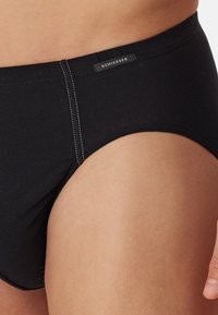 Zwarte katoenen slip met contrasterende stiksels en een label op de tailleband. De stof lijkt soepel met een strakke pasvorm rond het been.