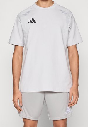 Homme portant un t-shirt de sport Adidas gris clair uni et un short assorti, debout avec les bras détendus le long du corps.