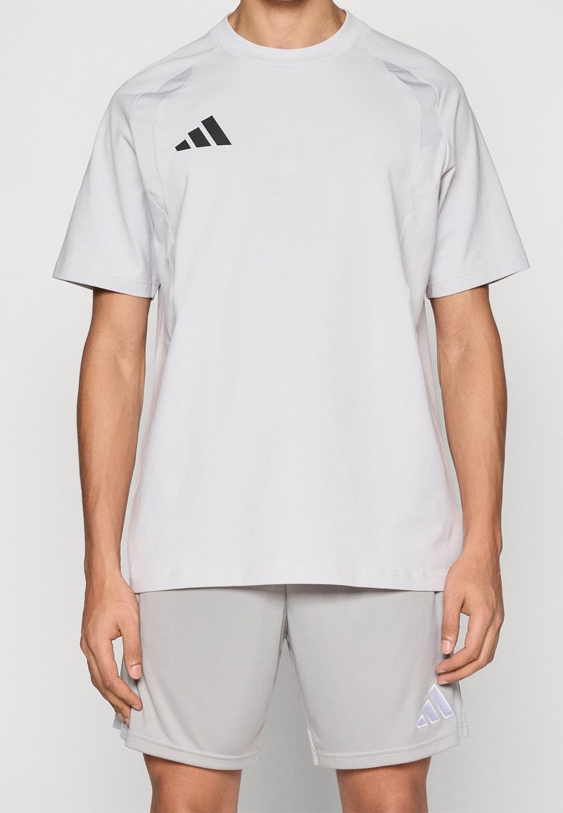 Homme portant un t-shirt de sport Adidas gris clair uni et un short assorti, debout avec les bras détendus le long du corps.