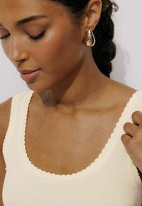 Débardeur couleur crème avec un décolleté en forme de vagues. Le modèle porte une boucle d'oreille en forme de goutte d'eau dorée et brillante. Le teint de la peau est chaud et lisse.