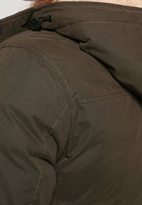 Close-up of a person wearing a brown padded jacket with visible seams and a black plastic toggle on the shoulder area.