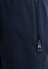 Jogging en tissu texturé bleu marine avec une poche zippée sur le côté. Présente un texte en relief et des accessoires de fermeture éclair contrastants.