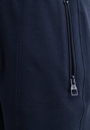 Tumši zila teksturēta auduma bikses ar sānu rāvējslēdzēja kabatu. Iespējas ar reljefa tekstu un kontrastējošu rāvējslēdzēju aparatūru.