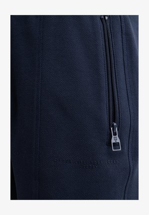 Tumši zila teksturēta auduma bikses ar sānu rāvējslēdzēja kabatu. Iespējas ar reljefa tekstu un kontrastējošu rāvējslēdzēju aparatūru.