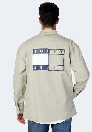 Mężczyzna w beżowej kurtce z dużym granatowo-białym logo Tommy Jeans na plecach, stojący na jednolitym jasnym tle.