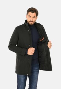 Cappotto isolato nero con colletto alto, zip frontale e bottoni a pressione. L'interno presenta un rivestimento trapuntato e una tasca con accento arancione.