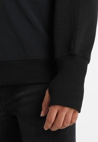 Felpa nera con polsini a coste, caratterizzata da una texture di tessuto liscia e un design senza cuciture sulle spalle. La mano è appoggiata su jeans neri.