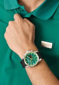 Orologio verde con quadrante testurizzato e dettagli in acciaio inox, cinturino in gomma e cassa in acciaio inox, abbinato a una polo verde.
