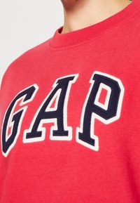 Czerwona bluza z okrągłym dekoltem, zdobiona haftowanym granatowym napisem "GAP" obramowanym na biało. Miękka, teksturowana tkanina, dopasowany krój.