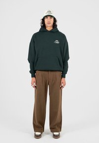 Donkergroene hoodie met een voorktas, gecombineerd met bruine plooibroek en schoenen met witte zolen, afgemaakt met een beige en groene pet.