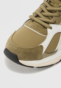 Olivenfargede sneakers laget av semsket skinn og nylon. Rund tå, hvite detaljer, og tykk svart såle. Snøring er stramt festet.