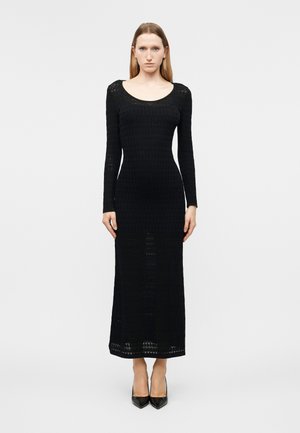 Moteris stovinti su atsipalaidavusiomis rankomis, vilkinti aptemptą juodą megztinį ilgomis rankovėmis ir juodus smailiais pirštais aukštakulnius ant vienspalvio fono.