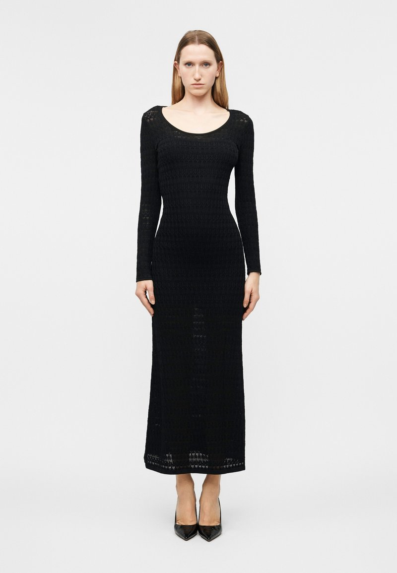 Γυναίκα σε όρθια στάση με χαλαρά τα χέρια, φοράει εφαρμοστό, μαύρο πλεκτό φόρεμα με μακριά μανίκια και μαύρες μυτερές ψηλοτάκουνες γόβες σε απλό φόντο.