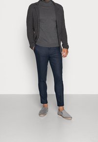 Muž v tmavě šedém roláku, strukturovaném tmavém kardiganu, námořnických slim kalhotách a světle šedých slip-on botách, stojí s rukama v kapsách.