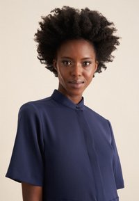 Abito camicia blu navy con maniche corte, colletto alto e chiusura con un singolo bottone. Tessuto liscio e design minimalista senza fantasie.