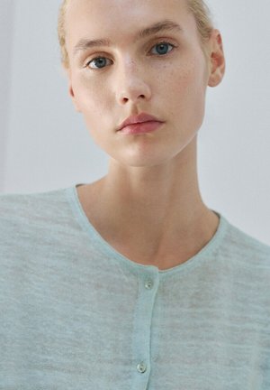 Giovane donna con pelle chiara, capelli biondi raccolti, che indossa una maglia azzurra con bottoni, che guarda direttamente con espressione neutra.