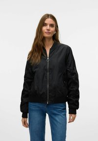 Veste bomber noire avec fermeture éclair sur le devant, poignets et ourlet côtelés, poches latérales et détails froncés sur les manches. Portée avec un jean bleu.