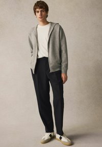 Un sweat à capuche zippé gris sur un t-shirt blanc, associé à un pantalon noir à rayures et des baskets beiges avec des accents noirs. Design et textures simples.