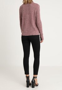 Vrouw die met haar rug naar de camera staat, gekleed in een roze gebreide trui, zwarte skinny jeans en zwarte hooggehakte enkelsandaaltjes.