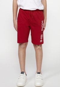 Rote Baumwollshorts mit elastischem Bund und verstellbarem Kordelzug. Mit bedrucktem Text an der Seite. Getragen mit weißen Sneakern.