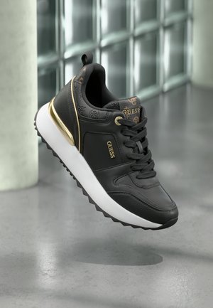 Zwarte sneaker van leer en textiel, met gouden accenten, vetersluiting, gestructureerd oppervlak en een witte zool met een gekartelde rand.