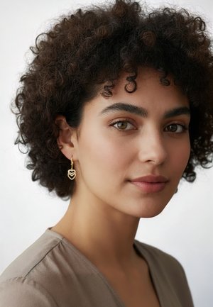 Guld hjerteformede øreringe med lagdelt design, båret af en person med krøllet mørkt hår, iført en bluse i neutrale nuancer mod en lys baggrund.