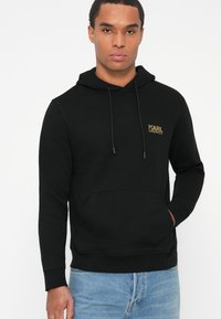 Joven usando una sudadera negra de Karl Lagerfeld con bolsillo canguro y jeans azul claro, de pie contra un fondo blanco liso.