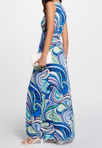 Maxi šaty bez rukávů s modrými, zelenými a fialovými spirálovitými vzory, doplněné bílými sandály a malou bílou kabelkou.