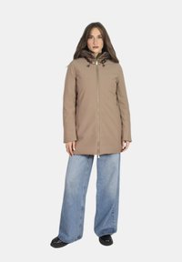 Giacca impermeabile marrone con zip frontale e cappuccio, dotata di foderatura interna lucida; indossata con jeans azzurri a gamba larga.