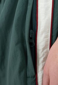 Nærbillede af en mørkegrøn jakke med en åben lynlås ved siden af en hvid og rød stofstribe, og en del af en hånd synlig til højre.