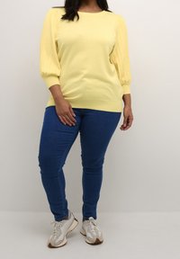 Gul stickad topp med ribbade puffärmar, kombinerad med blå skinny jeans och vita sneakers med ljusbruna detaljer.
