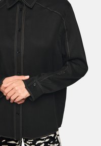 Black button-up shirt with contrasting beige stitching. Features a pointed collar, long sleeves, and black buttons along the front.